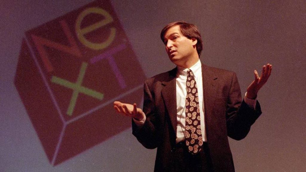 Man standing talking on a stage with arms outstreched and a logo behind him